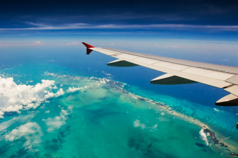 Aerial view from an airplane window showing a wing over turquoise tropical islands and coral reefs, highlighting the importance of having a travel camera ready during transit.