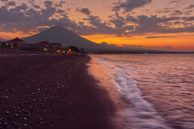 Sunset at a beach in Bali with Mount Agung in the background, silhouetted against a colorful sky reflecting on the water.